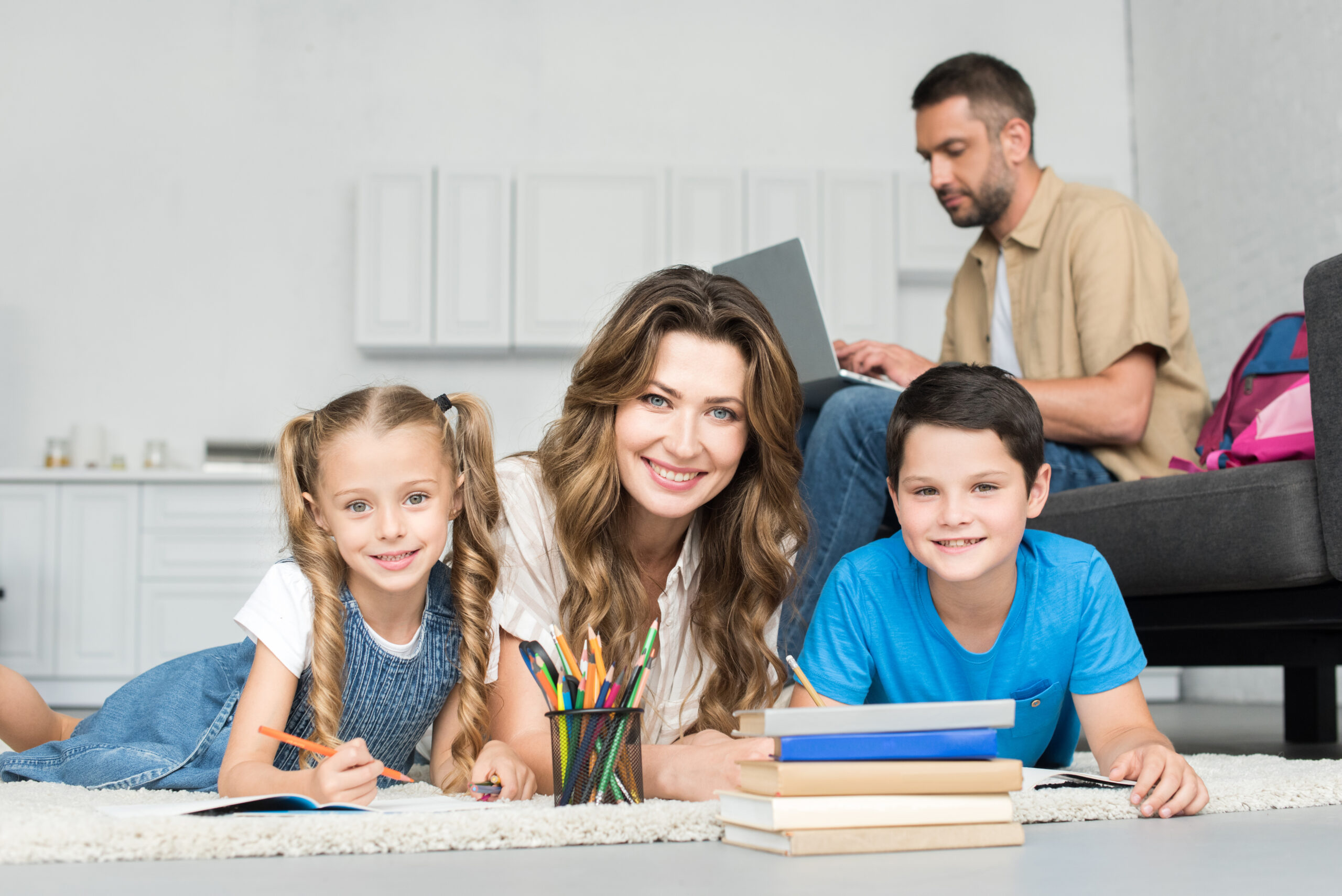 smiling mother and kids looking at camera while doing homework together and man using laptop on sofa