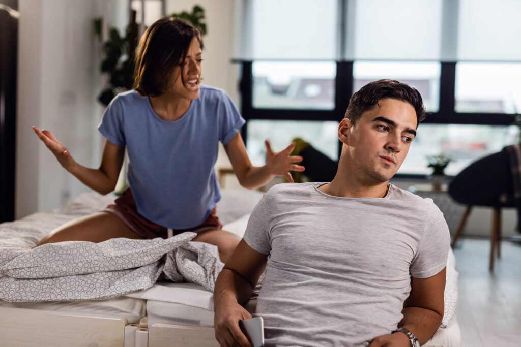 Young man feeling disconnected while sitting by the bed and ignoring his girlfriend during a fight.