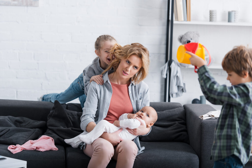 tired & stressed mother with a heavy mental load holding her infant kid sitting on sofa while naughty children playing at home